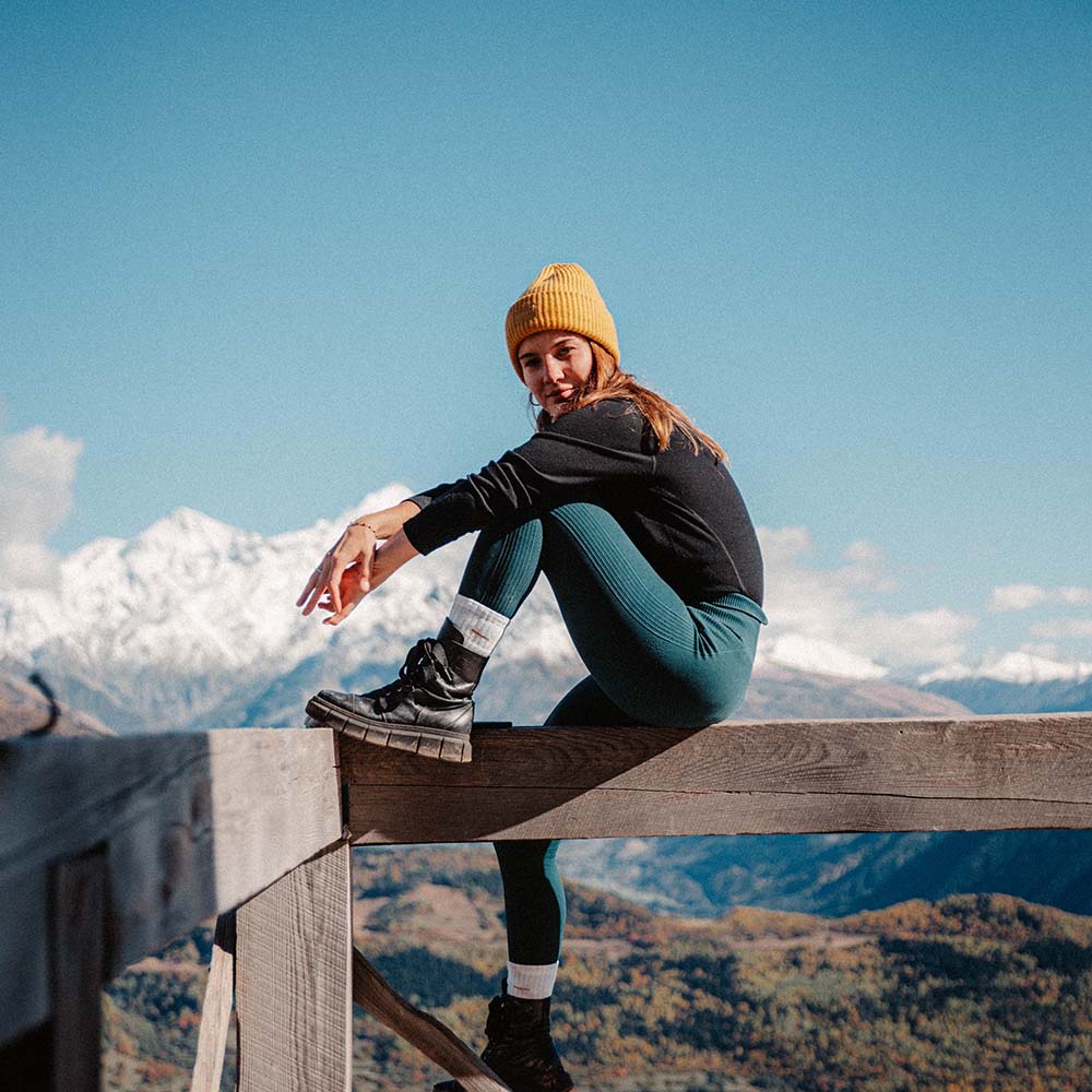 Frau trägt Merino Langarm Shirt Damen 230 g/m2 auf Bergzaun, umgeben von alpiner Landschaft. Perfekt für Outdoor-Abenteuer, bietet Komfort und Temperaturregulierung.