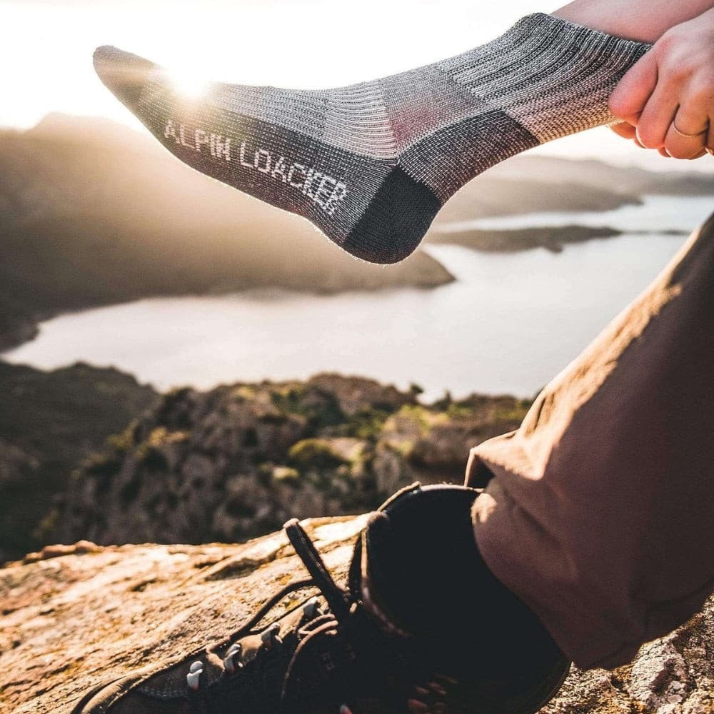 Hiker am Felsen mit Merino Wandersocken Classic Herren & Damen, ein Fuß in schwarzem Wanderschuh, der andere in grauer Socke, blickt auf sonnenbeschienenen See.