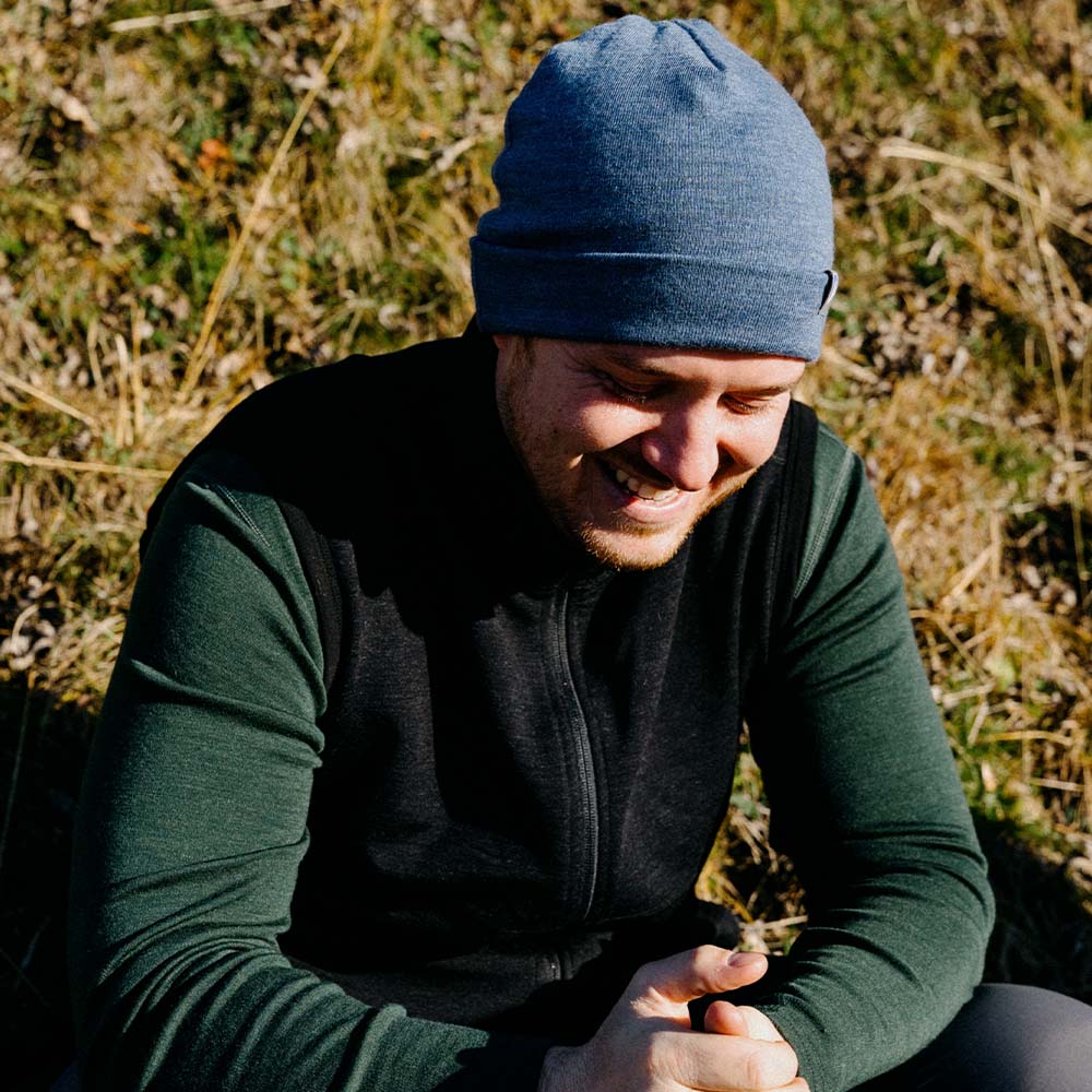Mann trägt Merino Weste Herren 270 g/m², lacht sitzend im Freien. Perfekt für Outdoor-Touren und Freizeit, bietet Komfort und Stil.
