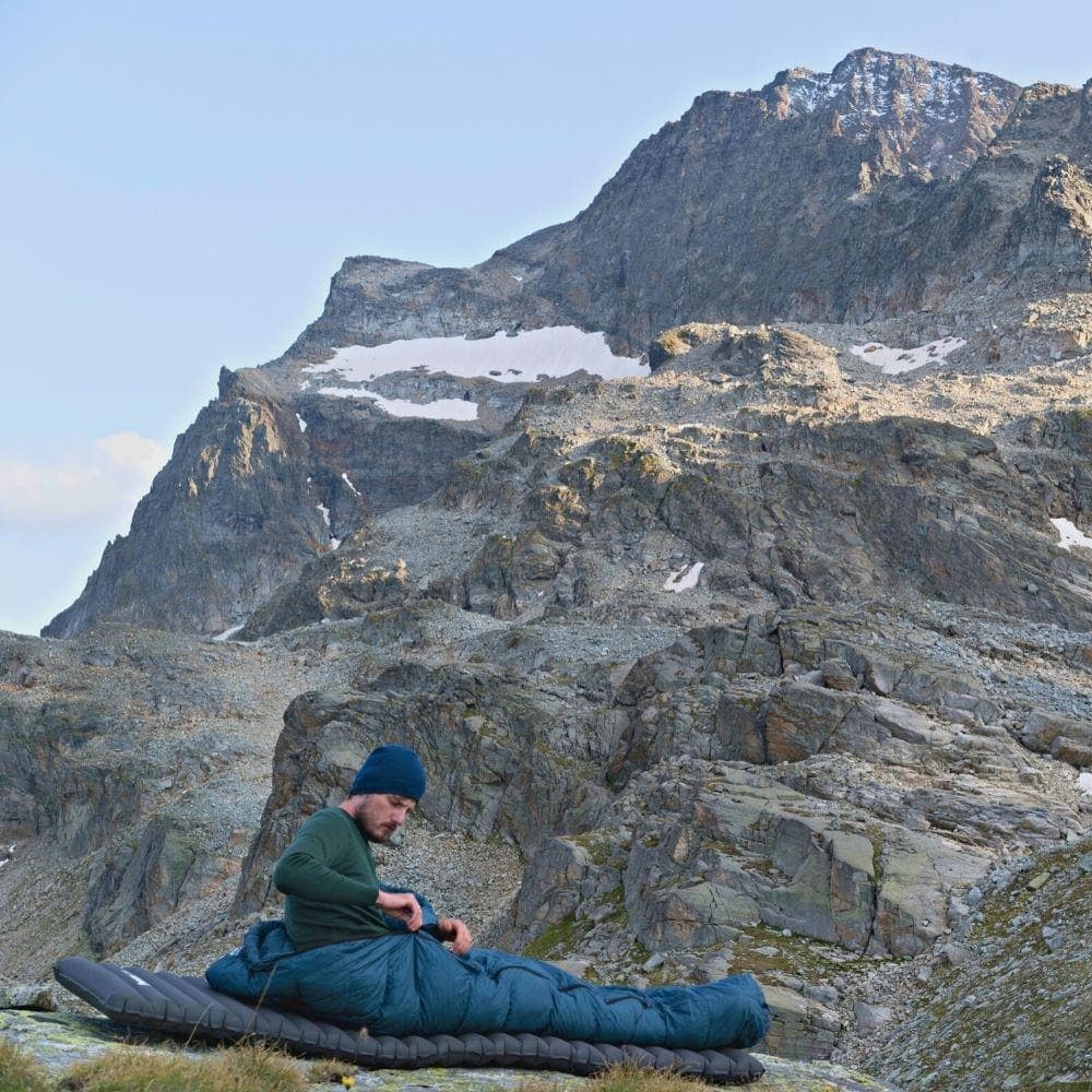 Person in Bergszene sitzt auf Schlafmatte im Down Pro 3 Jahreszeiten Schlafsack 860g, umgeben von Felsen und Schnee, ideal für Outdoor-Abenteuer.