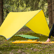 Campsite mit Tarp 500 als Regenschutz, darunter zwei Schlafmatten. Ein Wanderer entfaltet die gelbe Matte im Wald, ideal für Outdoor-Abenteuer.