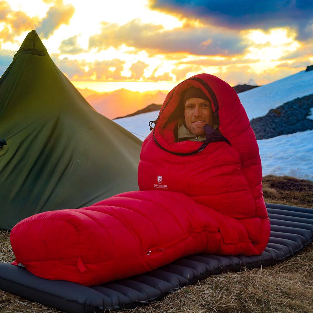 Person sitzt im Alpin Loacker Down Pro 4 Jahreszeiten Schlafsack vor Zelt in Berglandschaft bei Sonnenaufgang, ideal für kalte Outdoor-Abenteuer.