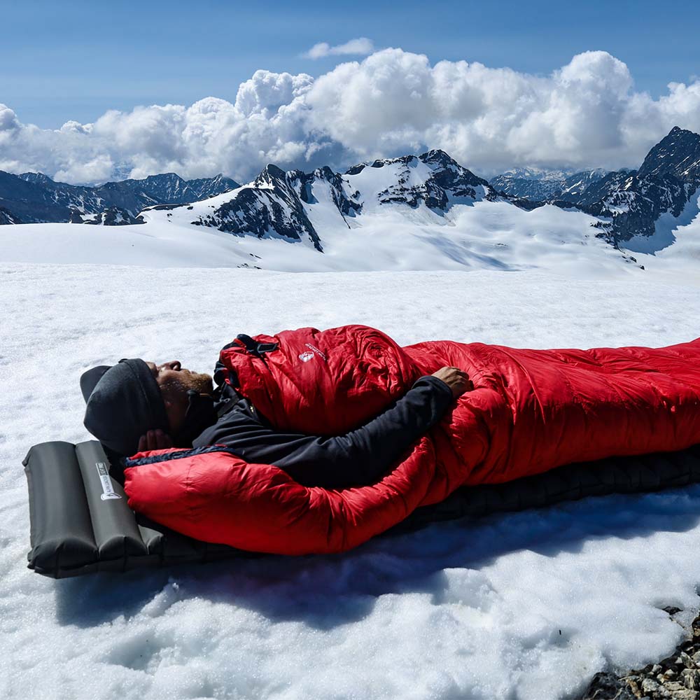 Person ruht in rotem Schlafsack auf der Leichte Isomatte isoliert mit Kunstdaune, auf schneebedecktem Gebirgskamm. Perfekt für alpines Camping und Trekking.