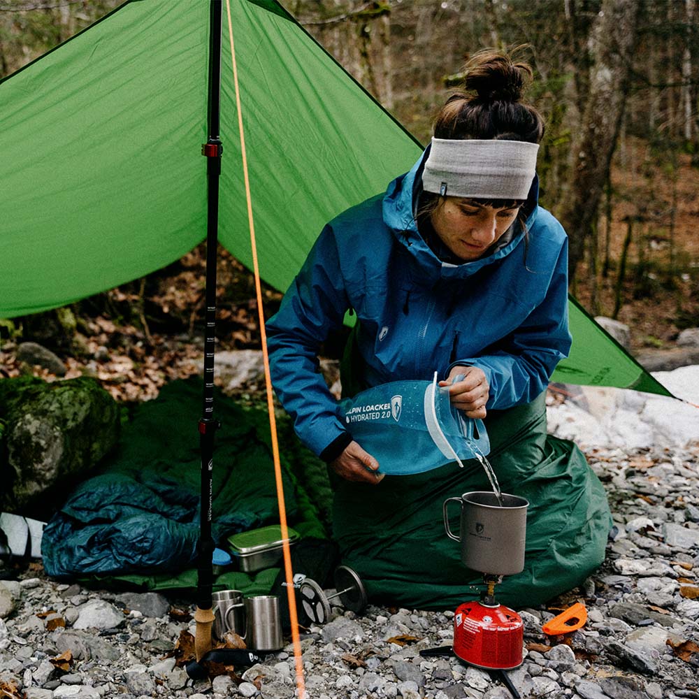 Person unter grünem Tarp 500 - das leichte Wingtarp, gießt Wasser in Campingbecher. Trekking-Stange stützt Tarp, daneben Schlafsack und Gaskocher auf Waldboden.