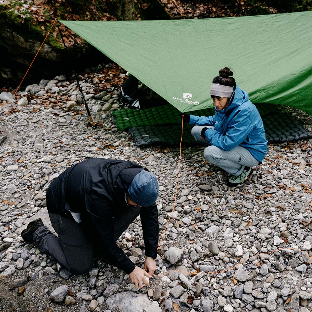 Tarp 500 - das leichte Wingtarp: Improvisiertes Lean-to mit Trekkingstöcken am Flussufer, ideal für Outdoor-Abenteuer. Person befestigt Abspannleine.