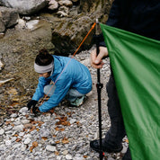 Person befestigt Tarp 500 am Flussufer, ideal für Outdoor-Abenteuer; vielseitig und leicht, bietet es Schutz bei Regen und Sonne für 1-2 Personen.