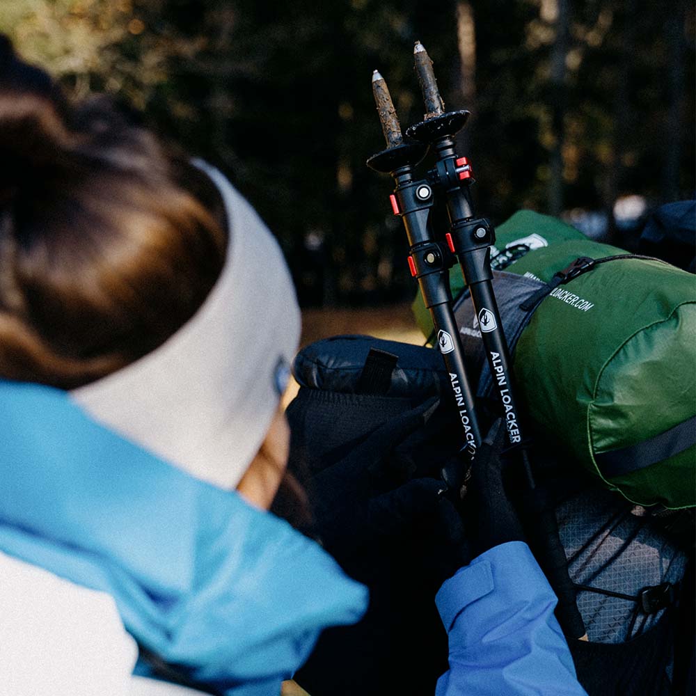 Hiker mit Alpin Loacker Teleskop Wanderstöcke aus Aluminium, befestigt am Rucksack im Wald; grüne Packtasche sichtbar. Ideal für Trekking- und Bergexpeditionen.