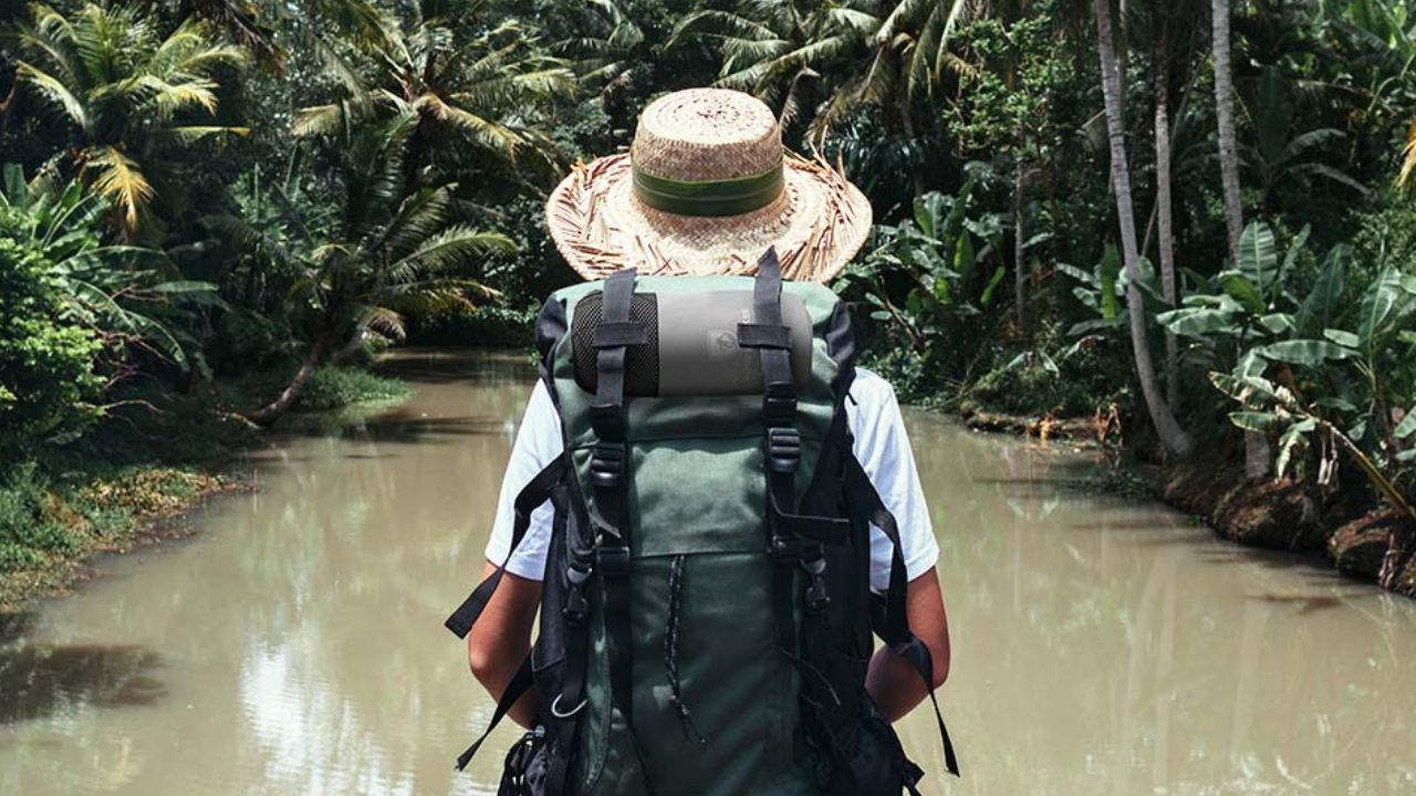 Eine Person mit Strohhut und Rucksack steht vor einem Gewässer, passend für Outdoor-Aktivitäten wie Wandern.