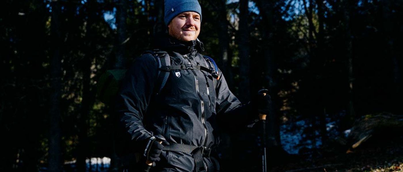 Ein Mann im schwarzen Anorak und Hut mit Rucksack im Wald, ideal für Outdoor-Abenteuer, wie sie bei alpinloacker.com angeboten werden.