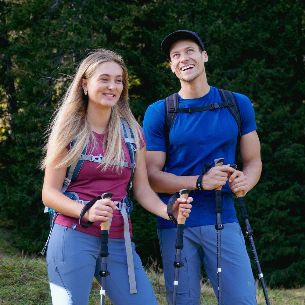 A man and a woman with hiking poles, both wearing outdoor clothing and backpacks, smiling outdoors, suitable for outdoor activities from alpinloacker.com.