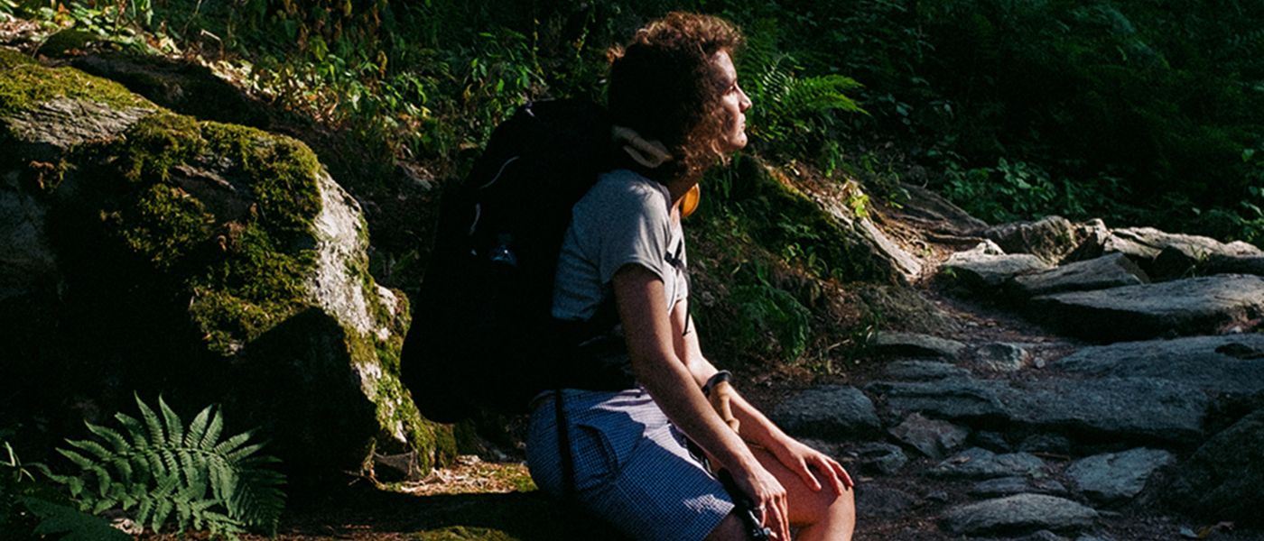 Person sitzt auf einem Felsen, trägt Outdoor-Kleidung. Umgebung deutet auf Wandern hin. Bild spiegelt Alpinloacker's Outdoor- und Wanderzubehör wider.
