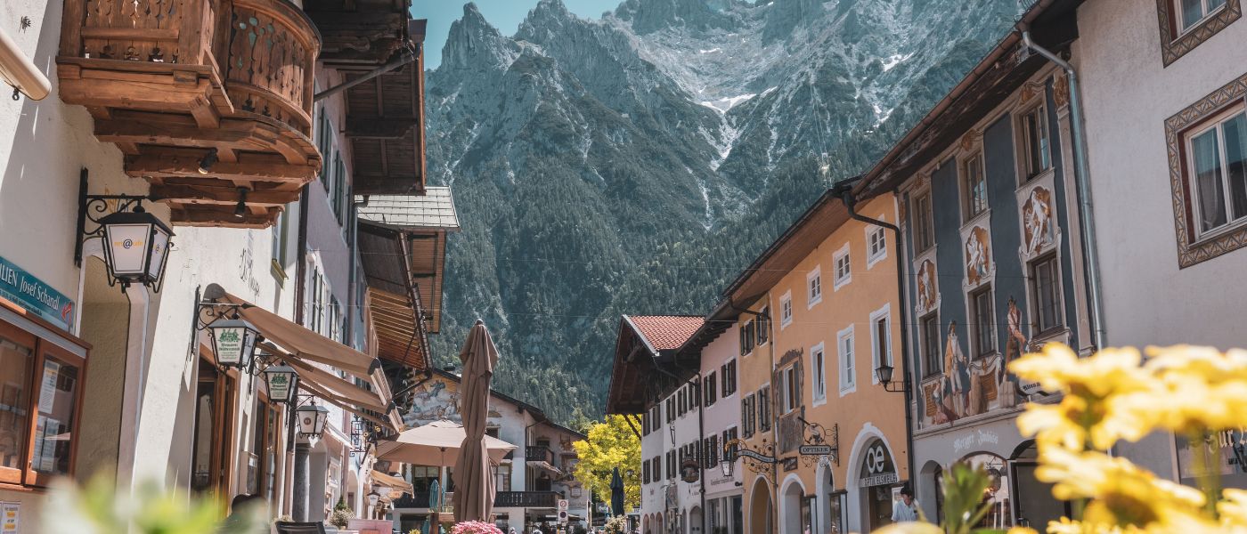 Straße mit Gebäuden und Bergen im Hintergrund, Nahaufnahme einer gelben Blume, passend zur Outdoor-Thematik von Alpinloacker's Sortiment für Wander- und Campingbedarf.