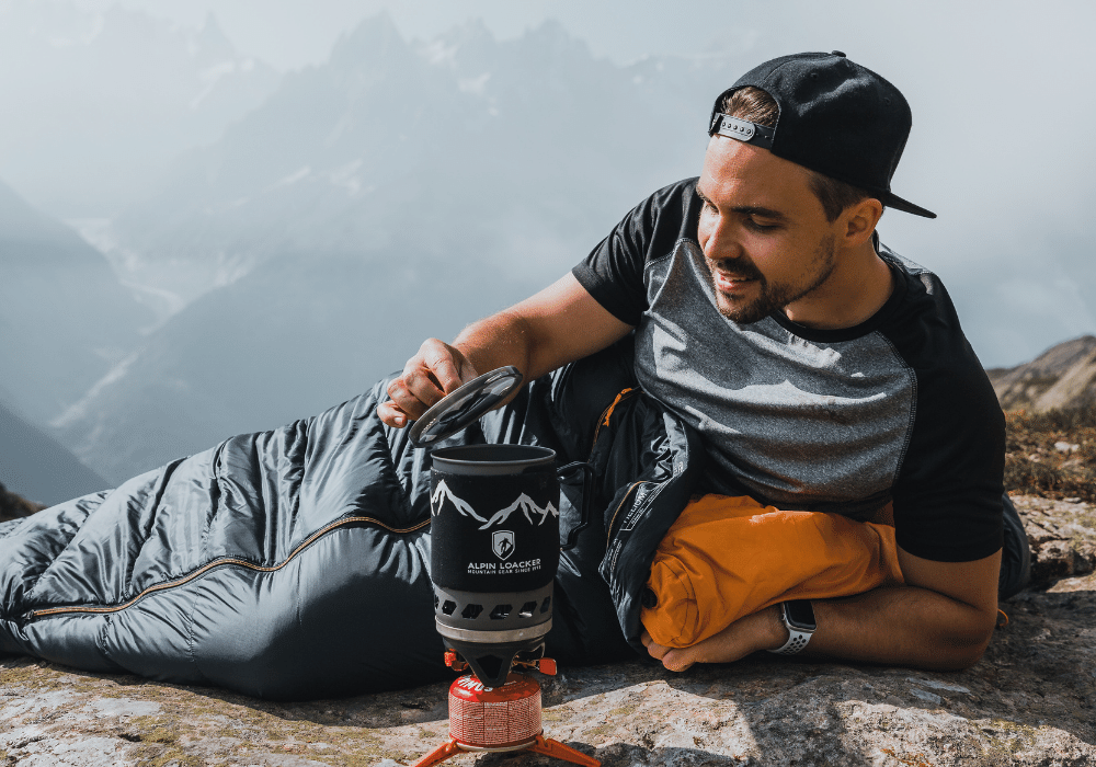 Ein Mann liegt auf einem Felsen neben einem Gaskocher, trägt Outdoor-Kleidung und blickt in die Natur. Ideal für Alpinloacker-Abenteuer.