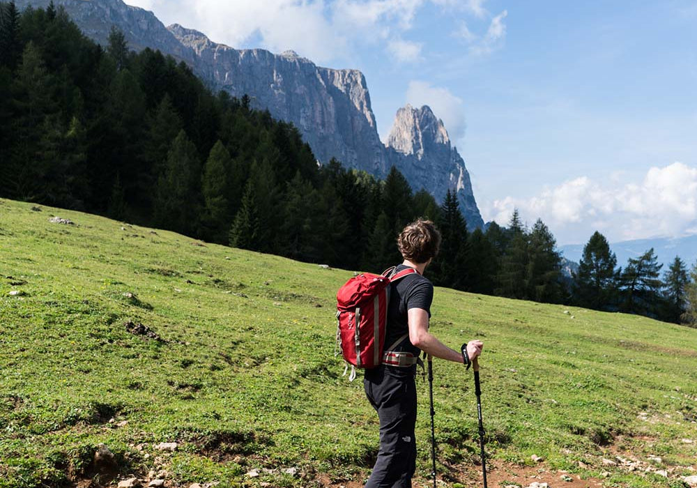 Ein Mann wandert mit Rucksack und Trekkingstöcken auf einem grasbewachsenen Hügel vor einem bewölkten Himmel.