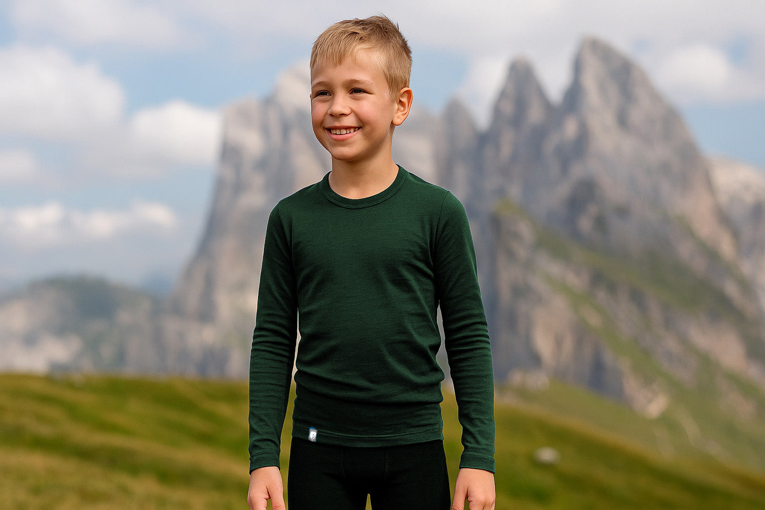 Junger Junge im Freien in einer Bergwiese, trägt dunkelgrünes Langarmshirt und schwarze Hose, lächelnd vor grasigen Hängen und felsigen Gipfeln unter bewölktem Himmel.
