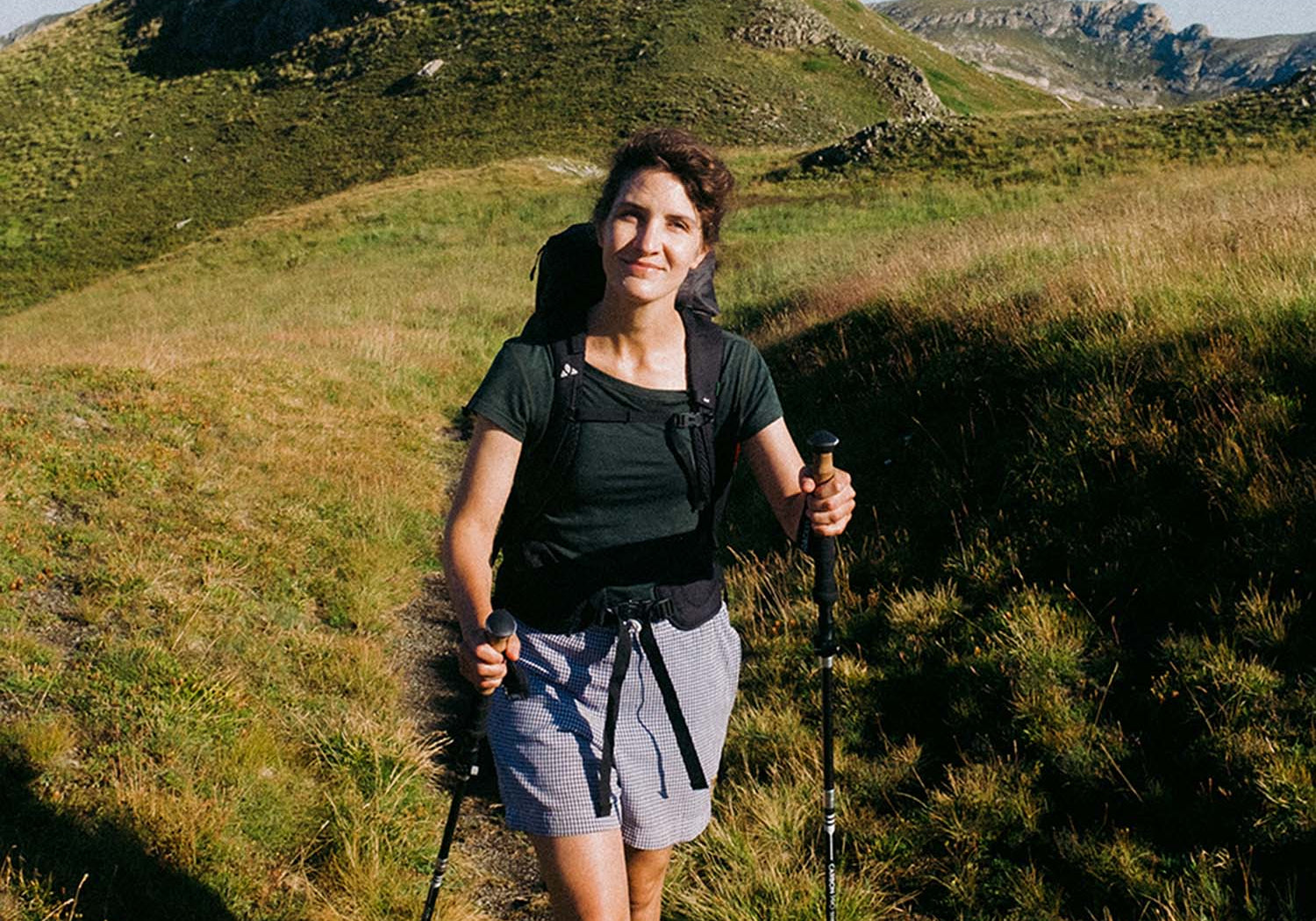 Eine Frau wandert mit Rucksack und Wanderstöcken im Gras, passend zu Outdoor-Ausrüstung von alpinloacker.com, spezialisiert auf leichte Wanderausrüstung.