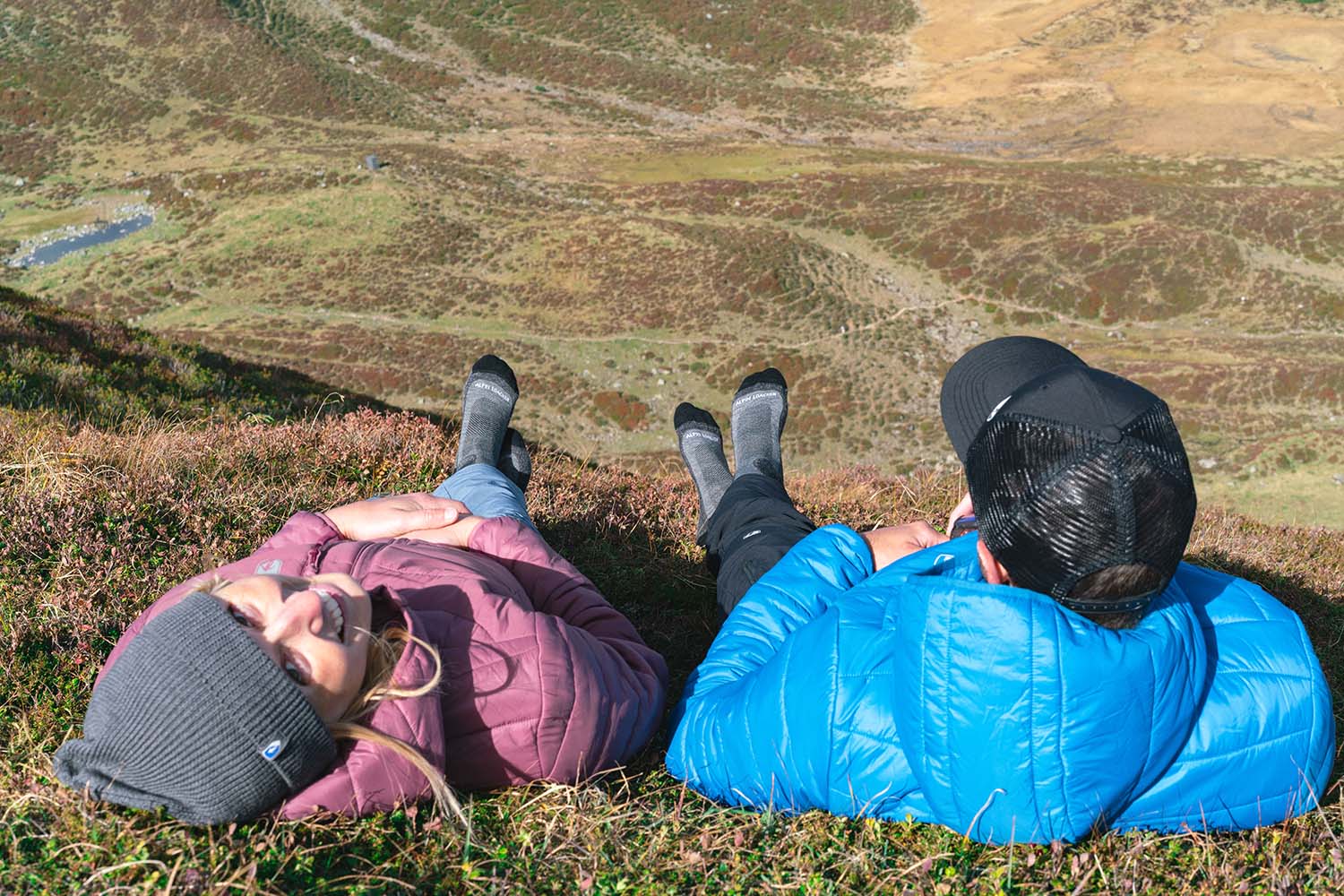 Ein Mann und eine Frau liegen lächelnd auf einer Wiese, beide tragen Outdoor-Kleidung, passend zum Thema Wandern und Naturerlebnis von Alpinloacker.