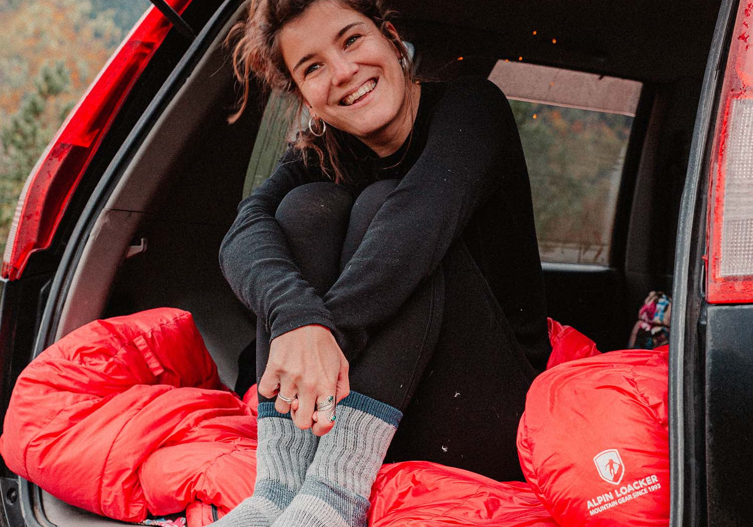 Eine Frau sitzt lächelnd im Auto, trägt graue Socken und hält eine rote Tasche mit Logo.