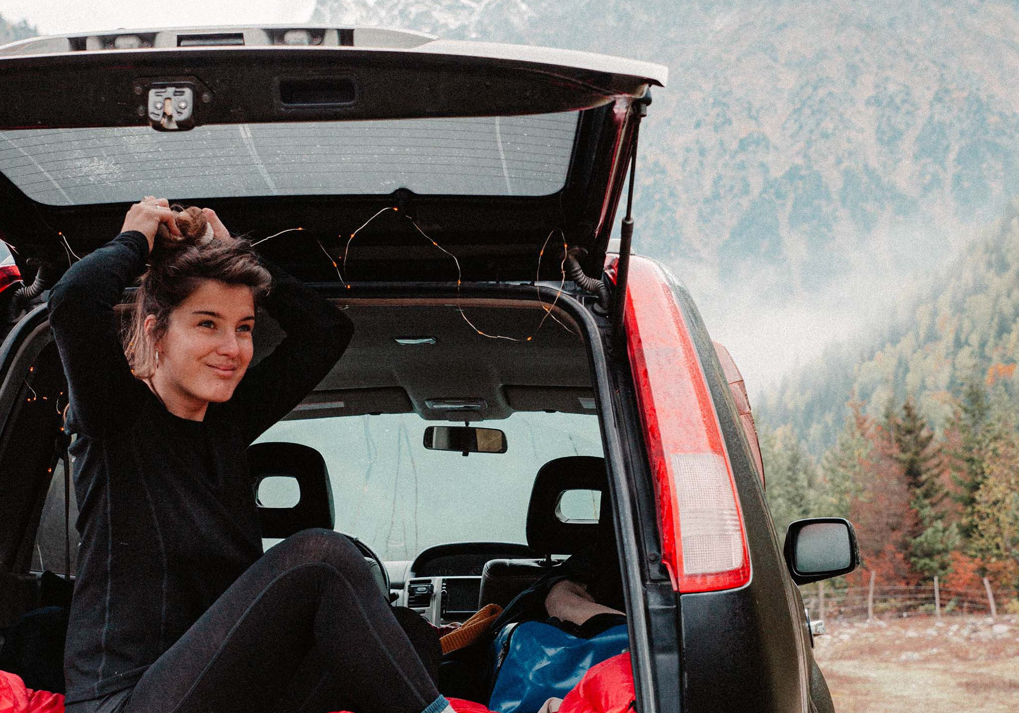 Eine Frau sitzt im Fond eines Autos, umgeben von Outdoor-Ausrüstung, passend für Aktivitäten wie Wandern und Camping.