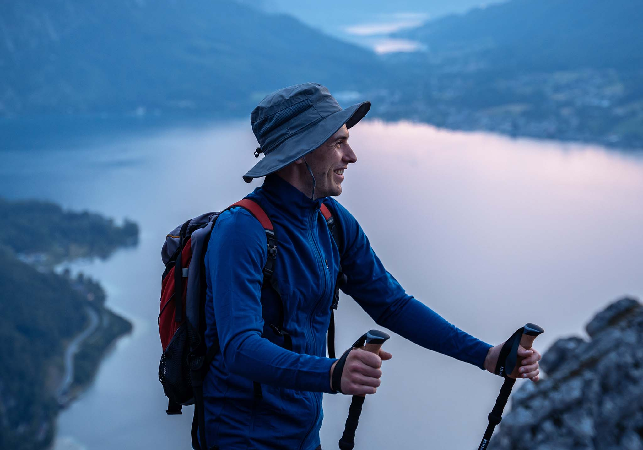 Ein Mann mit Hut und Rucksack wandert mit Stöcken auf einem Berg, im Hintergrund ein See und Berge.