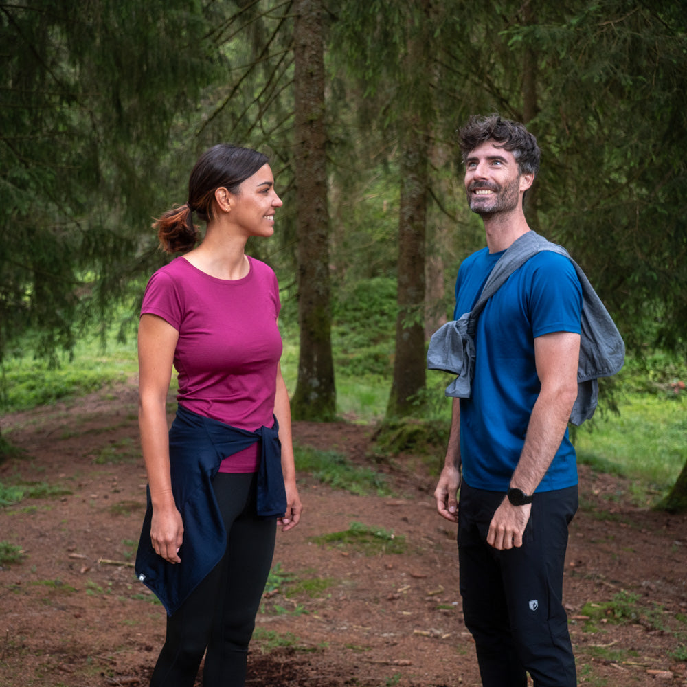 Zwei Personen in Wanderkleidung auf Waldweg: Frau in magentafarbenem Top, Mann in blauem T-Shirt, umgeben von hohen Nadelbäumen.