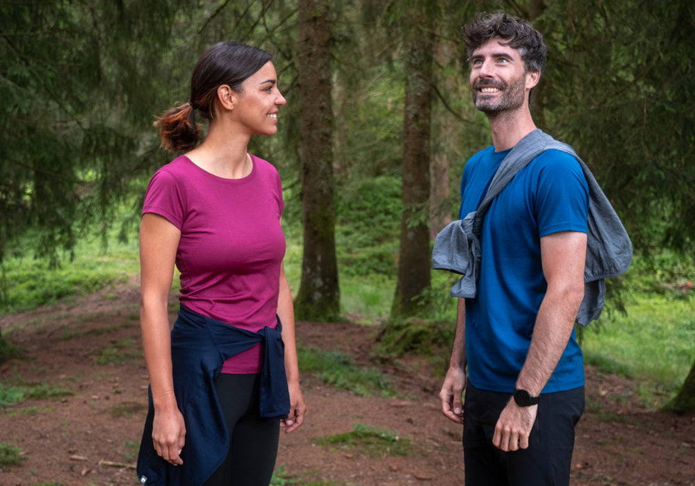 Zwei Personen in Wanderkleidung auf Waldweg: Frau in magentafarbenem Top, Mann in blauem T-Shirt, umgeben von hohen Nadelbäumen.
