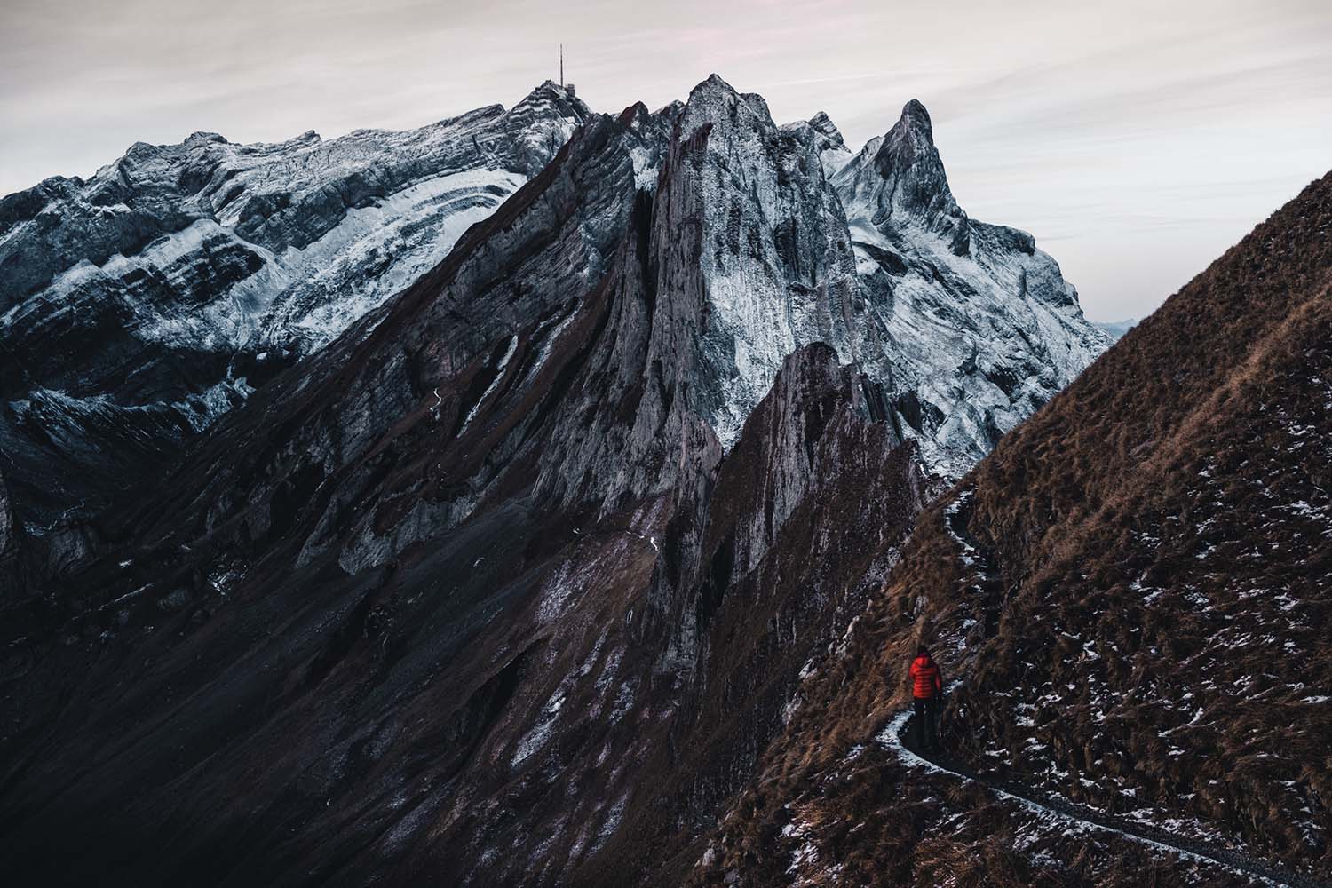 A person in a red and black striped shirt walks along a snow-covered mountain path, surrounded by an impressive mountain panorama.