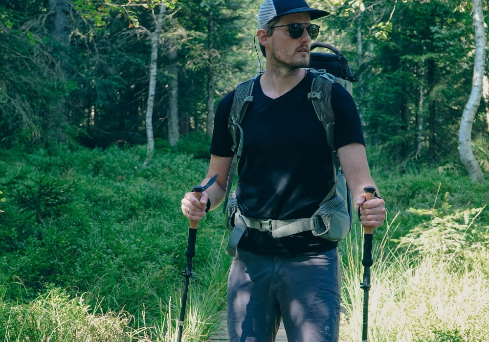 Ein Mann mit Rucksack und Sonnenbrille wandert mit Stöcken durch einen Waldweg, symbolisiert Outdoor-Ausrüstung und Wanderfreude.