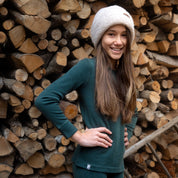 Ein Mädchen steht in 100% Merino langarm Set in grün vor einem Holzstapel