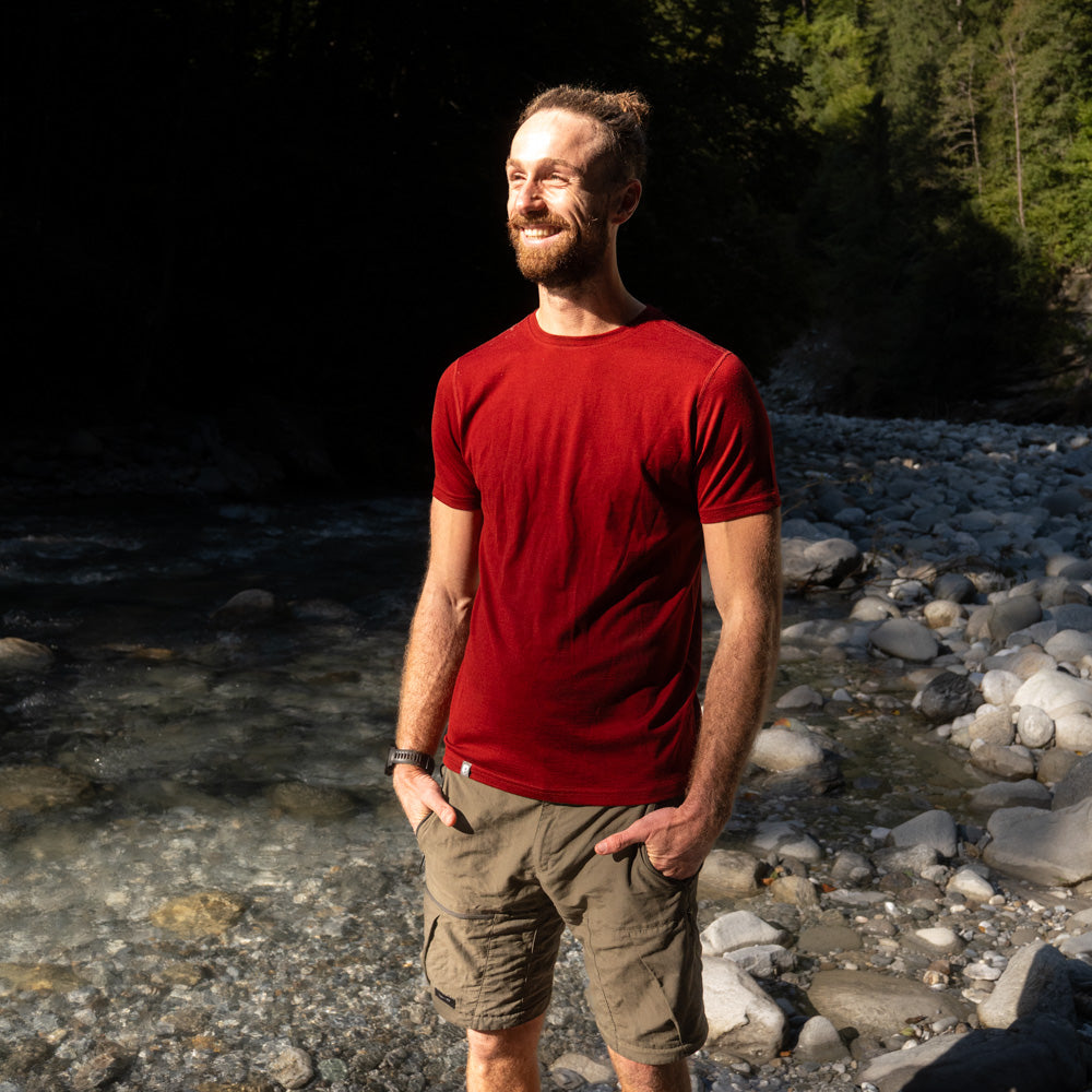 Mann steht lächelnd in der Natur und trägt kurzärmliges, rotes 180g Merino Shirt - perfekt für jede Outdoor Aktivität