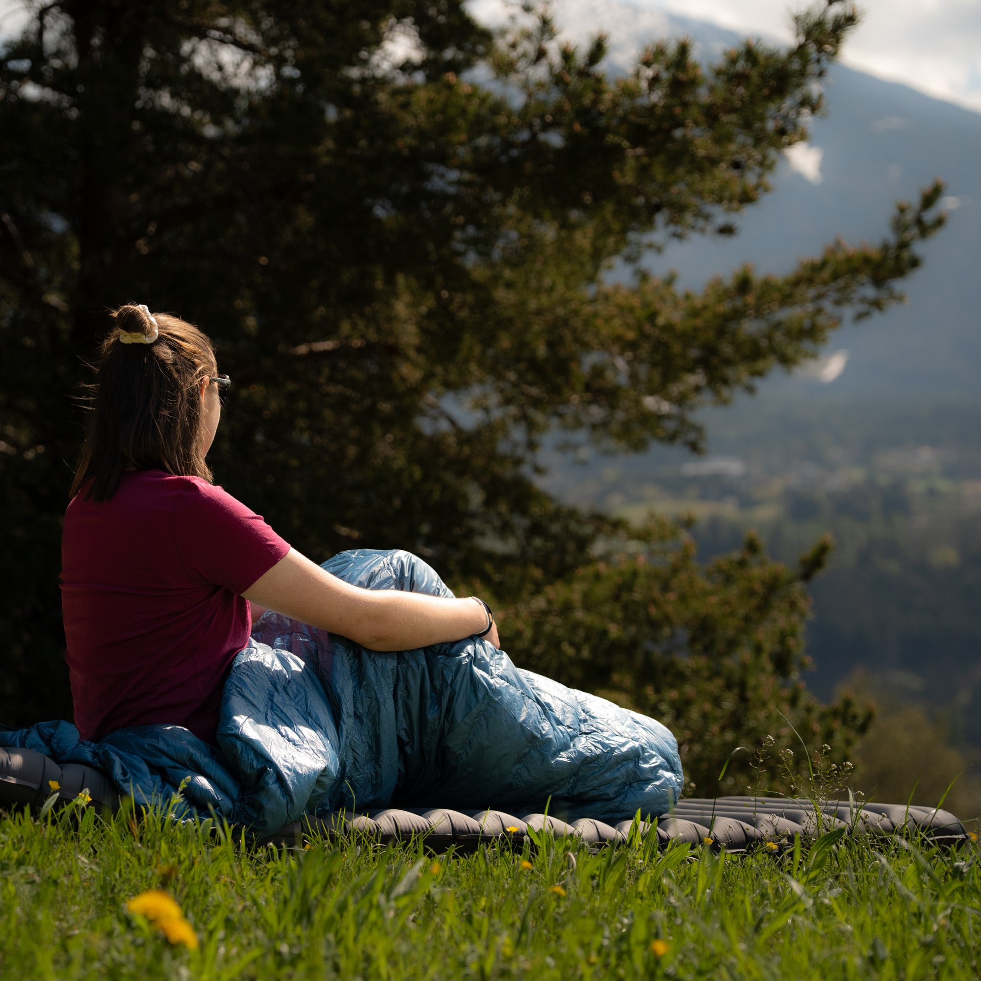 Model sitzt in blauem Sommerdaunenschlafsack auf der Wiese und schaut in die Ferne