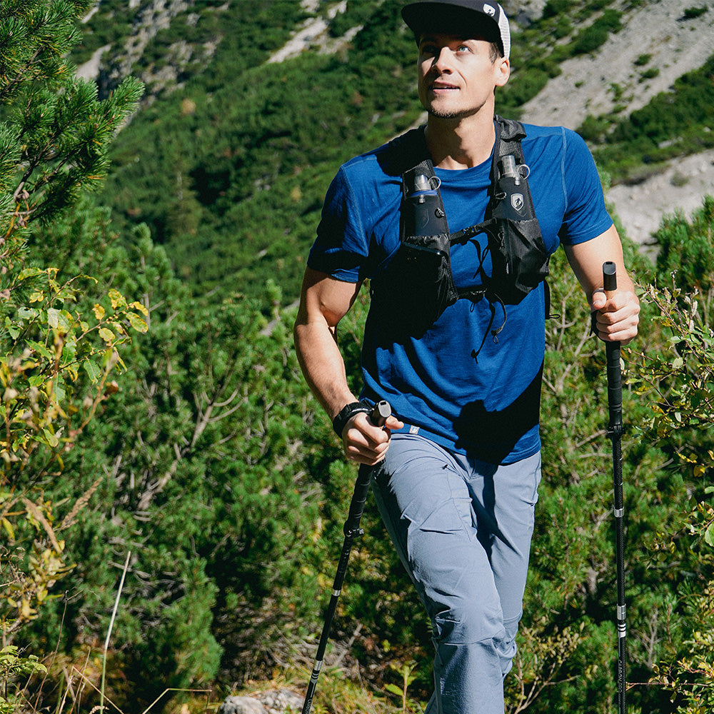 Gilet de course Trail Pro 5 avec 2 bouteilles, porté par un homme lors d'une randonnée en montagne avec des bâtons, idéal pour un trail running léger et fonctionnel.