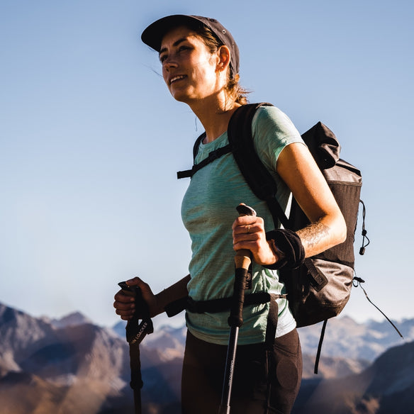 A woman with a rucksack and hiking poles wears the 150g/m2 women's merino T-shirt, ideal for outdoor activities thanks to temperature regulation and moisture wicking.