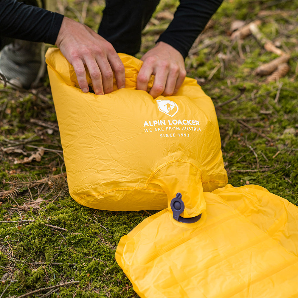 Hände öffnen eine gelbe Tasche der ultraleichten Isomatte, isoliert mit Kunstdaune, ideal für Outdoor-Abenteuer.