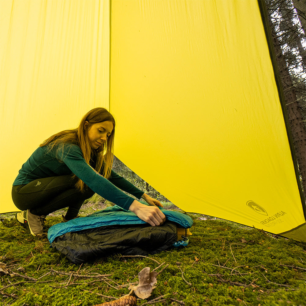 Frau kniet und bindet das Tarp 500 - das leichte Wingtarp, ideal für Outdoor-Abenteuer und Schutz vor Regen und Sonne, zusammen.