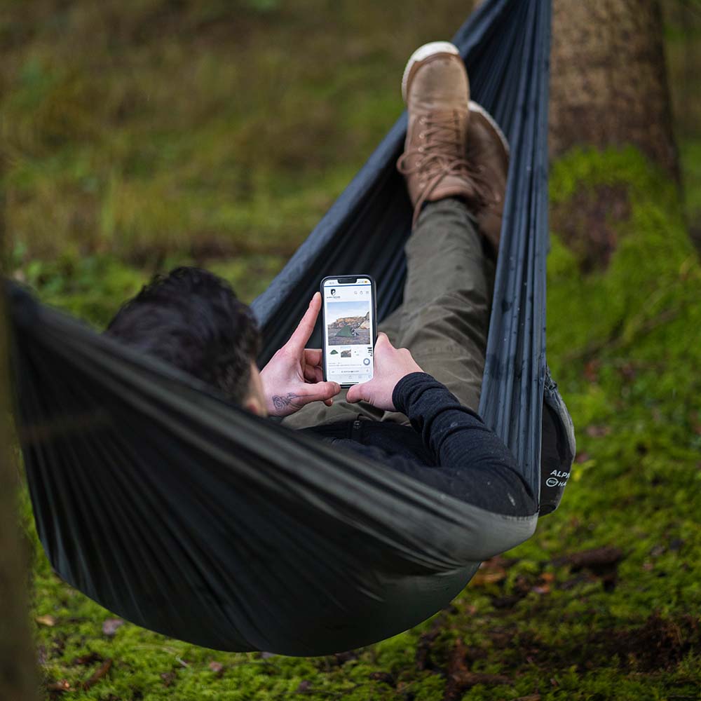 Hombre en el bosque en una hamaca ligera gris negra Alpin Loacker para senderismo y acampada al aire libre, hamaca con mosquetones y mochila