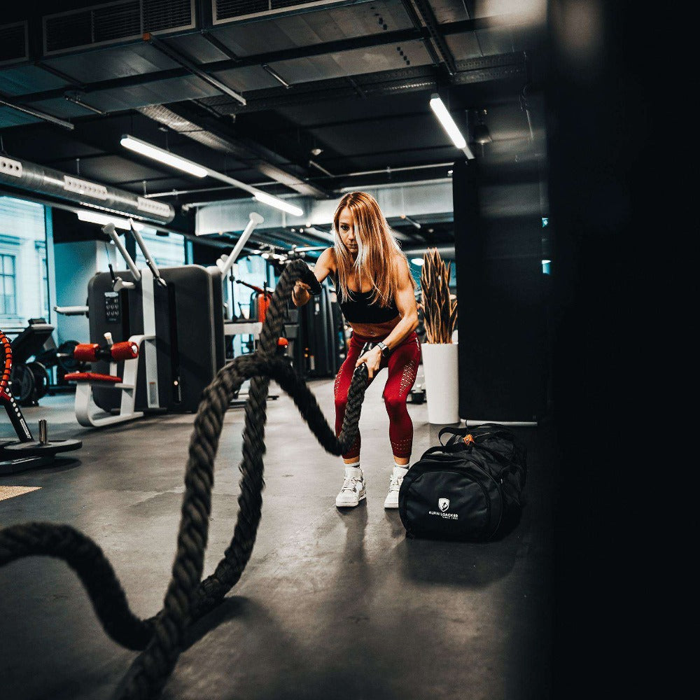 Frau im Fitnessstudio neben der Reisetasche, Sporttasche & Duffel Bag von Alpin Loacker, ideal für Abenteuer, mit PVC-Material und mehreren Fächern.